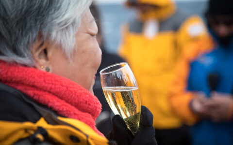 Woman in yellow parka & red neck gaiter holds glass of champagne while toasting crossing the Antarctic Circle on an expedition.