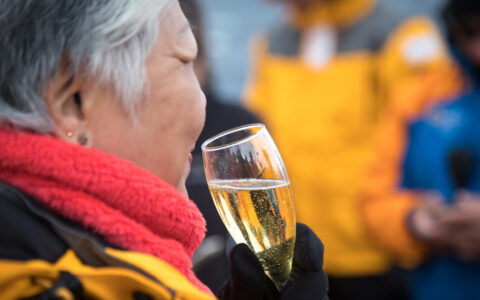 Woman in yellow parka & red neck gaiter holds glass of champagne while toasting crossing the Antarctic Circle on an expedition.