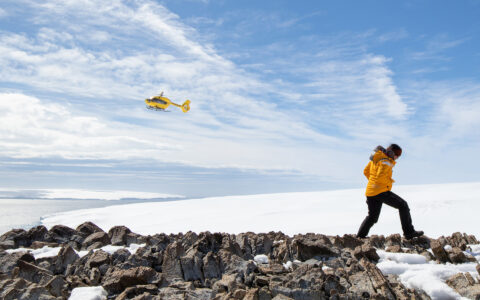 Antarctic Circle traveler in yellow parka walks over rocky peak with snowfield beyond & yellow helicopter flying overhead.