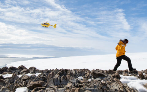 Antarctic Circle traveler in yellow parka walks over rocky peak with snowfield beyond & yellow helicopter flying overhead.