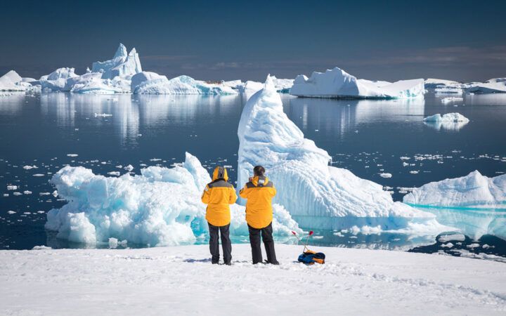 Two travelers in yellow polar parkas stand on snow gazing over white icebergs in blue water on an Antarctic trip.
