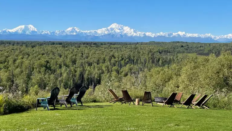 Large green lawn with Adirondack chairs around a firepit overlooking forest and snowcapped Mt. Denali in the distance.
