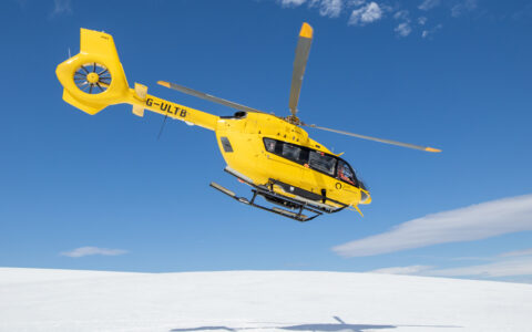 A bright yellow helicopter takes flight off the snow covered mountains