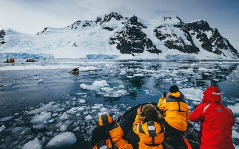 Antarctica travelers in bright jackets sit in Zodiac boats & photograph an orca coming out of the icy water.