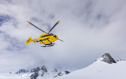 A bright yellow helicopter takes flight off the snow covered mountains