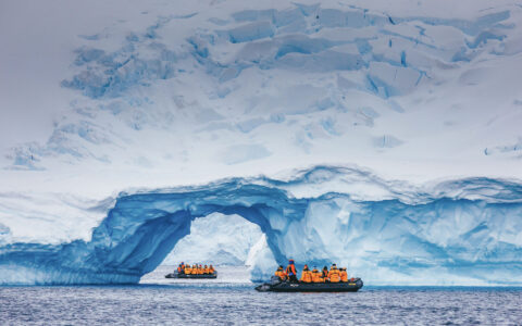 2 zodiacs with Antarctica travelers in bright jackets cruise amongst a snowy arch during an emperor penguin expedition.