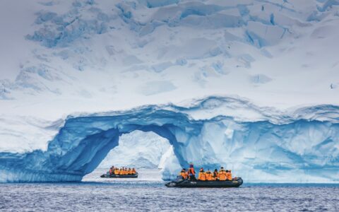 2 zodiacs with Antarctica travelers in bright jackets cruise amongst a snowy arch during an emperor penguin expedition.