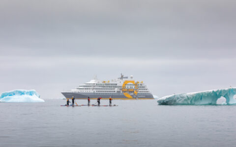 paddle boarders braving the icy cold waters next to two large icebergs in front of Quark expedition cruise ship