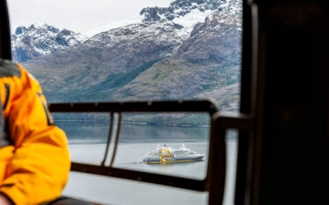 Chile cruise guest in yellow jacket sits in helicopter looking at Patagonia's peaks & Ultramarine expedition ship down below.