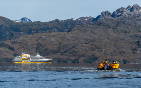 Zodiac with Chile cruise travelers makes its way through calm water toward Ultramarine expedition ship beside jagged peaks.