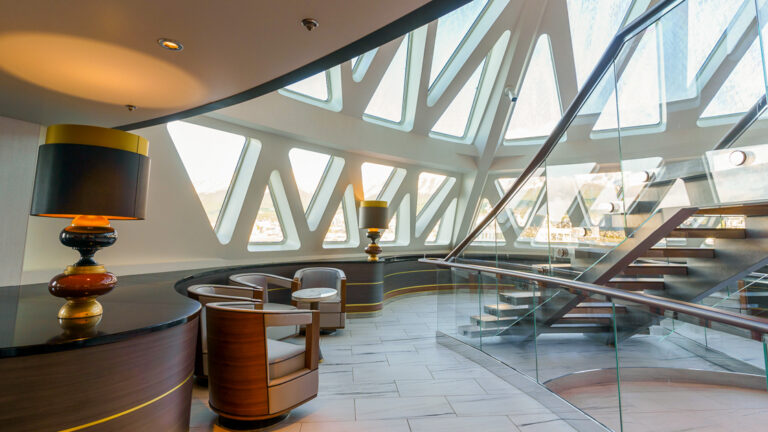 Glass atrium with triangular windows, white & wood captain's chairs, tile floor & large staircase on Ocean Explorer ship.