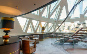 Glass atrium with triangular windows, white & wood captain's chairs, tile floor & large staircase on Ocean Explorer ship.