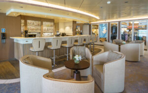 Latitude Bar on Ocean Explorer polar ship in gray & white with wood & stone decor, backed barstools & lounge chairs behind.