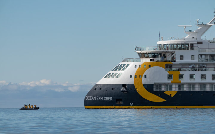 Zodiac boat approaches slim, pointed bow of Ocean Explorer expedition ship with gray, yellow & white hull & white upper decks & many windows.