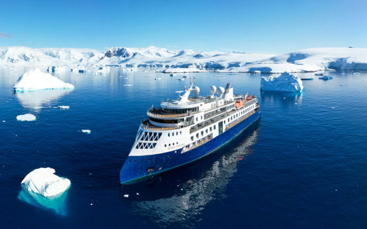 Aerial view of Ocean Explorer expedition ship in icy water with blue hull & white upper decks, slim bow, many windows & teak decks.