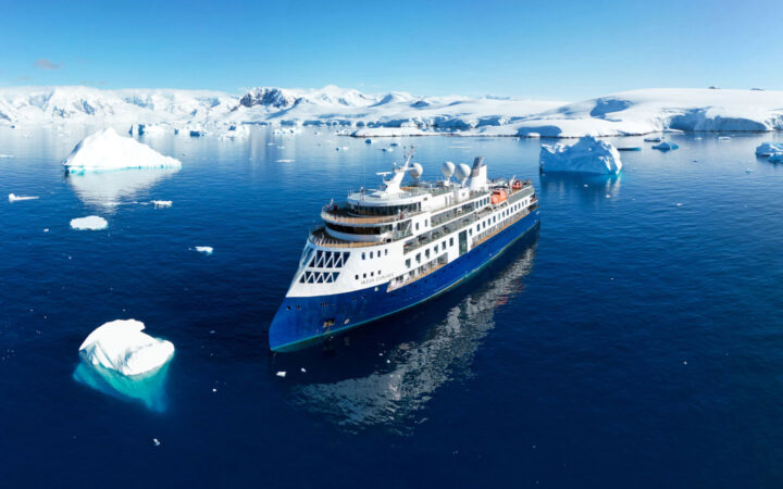 Aerial view of Ocean Explorer expedition ship in icy water with blue hull & white upper decks, slim bow, many windows & teak decks.
