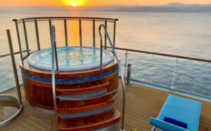 Round Jacuzzi with glass panels on one corner & wood & tile exterior sits atop teak deck of Ocean Explorer ship at sunset.