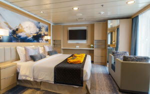 Owner's suite bedroom on Ocean Explorer with double bed in white sheets & gray couch by floor-to-ceiling windows & big balcony.