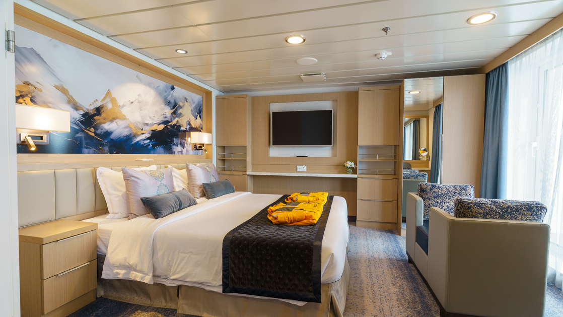Owner's suite bedroom on Ocean Explorer with double bed in white sheets & gray couch by floor-to-ceiling windows & big balcony.