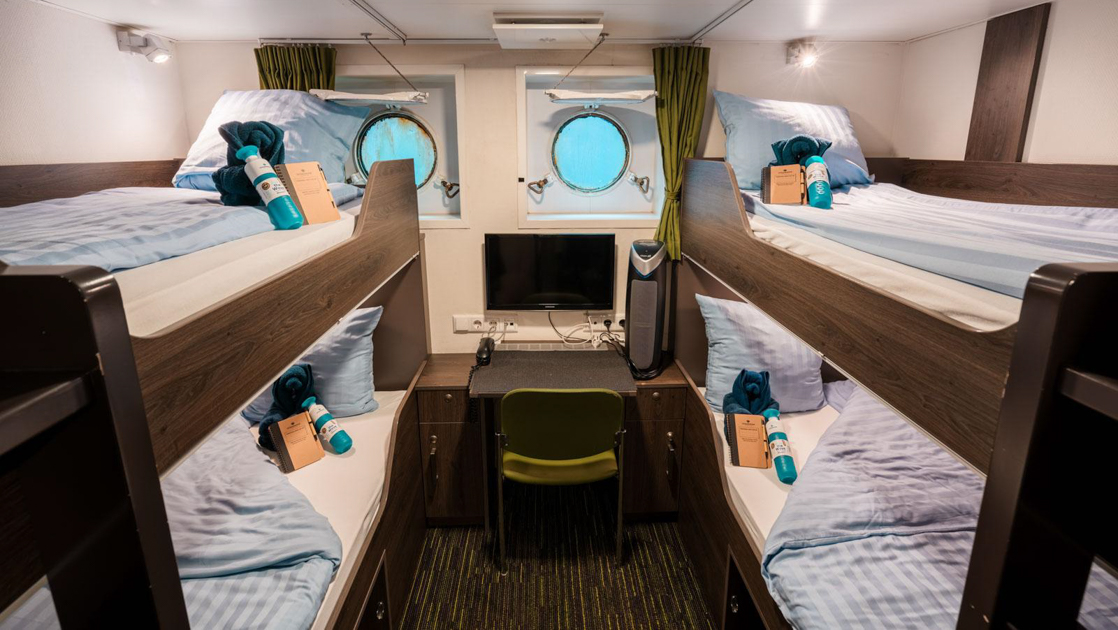 Ortelius small ship cabin with 2 sets of bunkbeds separated by a small desk with TV below 2 small portholes.