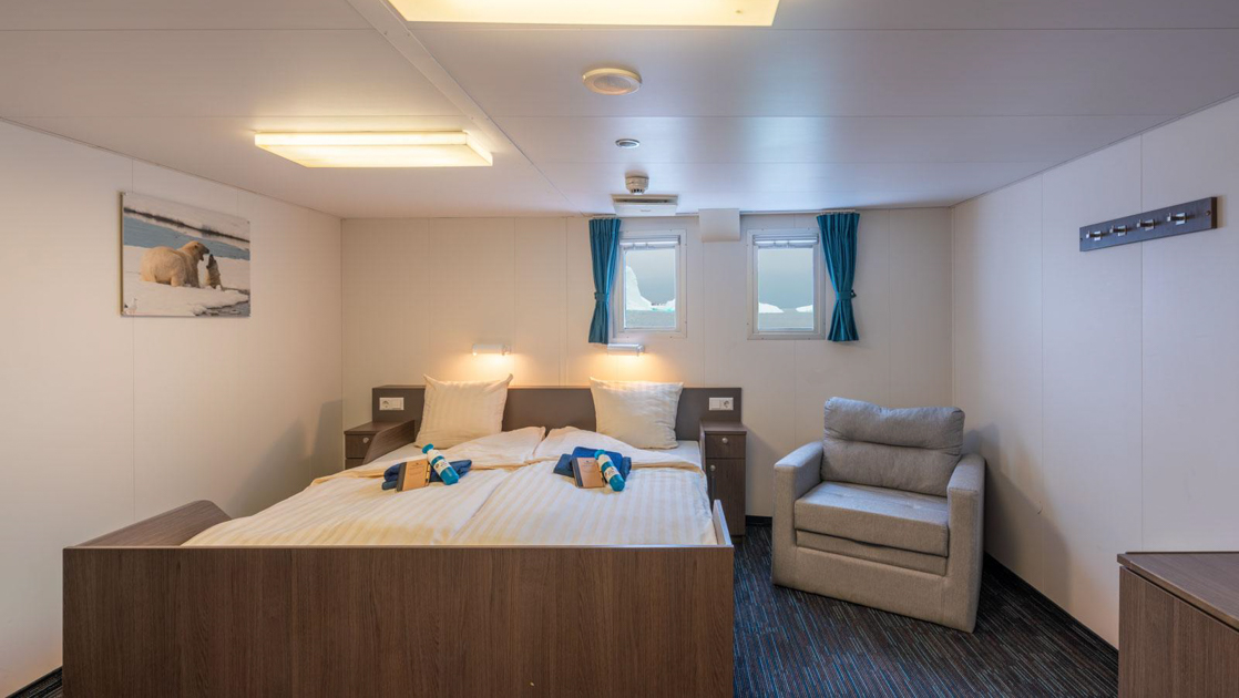 Superior cabin on Ortelius ship with double bed & gray chair, wood desk & white walls with polar bear photo & wall hooks.