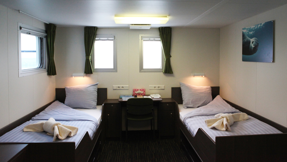 Twin Deluxe cabin on Ortelius ship with 2 beds in white linens with desk between & 3 small windows among white walls.