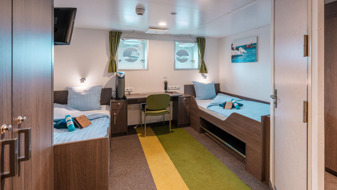 Twin porthole cabin aboard Ortelius ship with 2 twin-size beds in white linens, separated by a desk, plus 2 portholes & armoire.