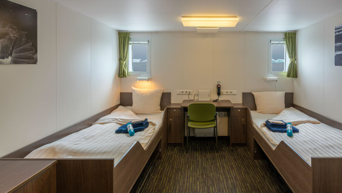 Ortelius ship Twin Window cabin with 2 twin-size beds in white linens separated by small desk & 2 small view windows.