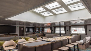 Library on Oretlius ship with bookcase shelves filled with books by view windows under bright skylight ceiling.