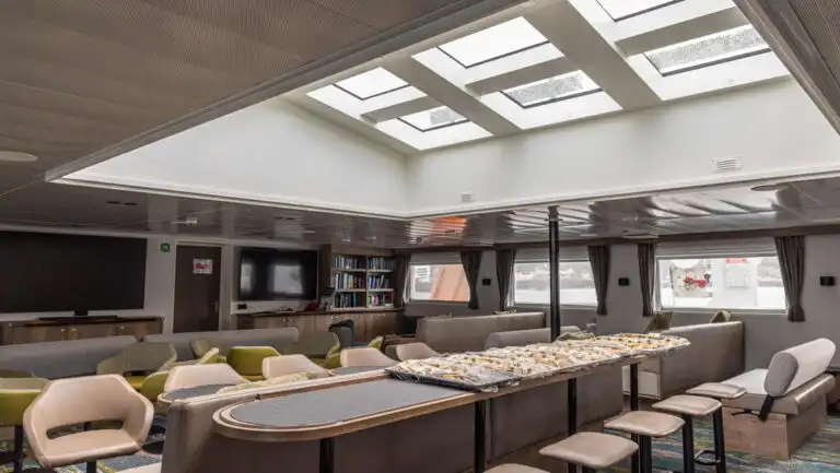 Library on Oretlius ship with bookcase shelves filled with books by view windows under bright skylight ceiling.