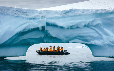 Zodiac passengers cruise through a dramatic blue iceberg arch in Antarctica.