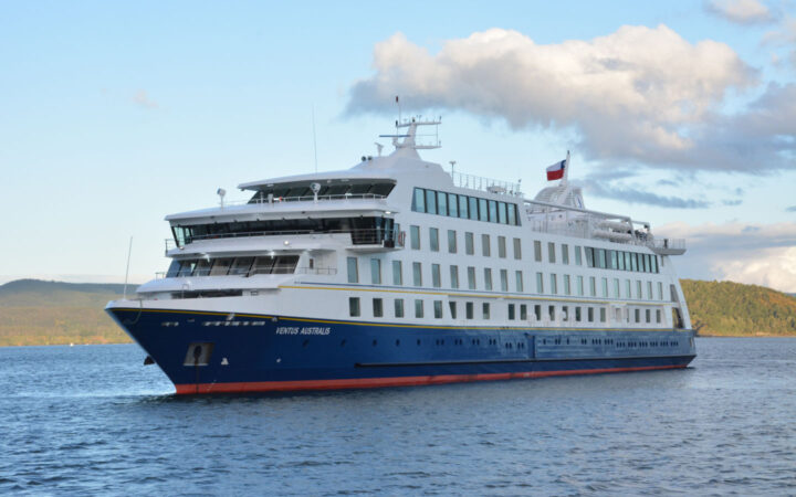 Ventus Australis expedition ship with blue hull & 4 white upper decks, cruising in calm sea with a Chilean flag flying.