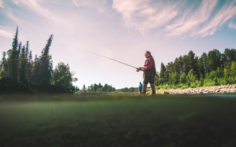 Woman stands at river's edge fishing while nearby man watches on a sunny day of the Within the Wild Alaska tour.
