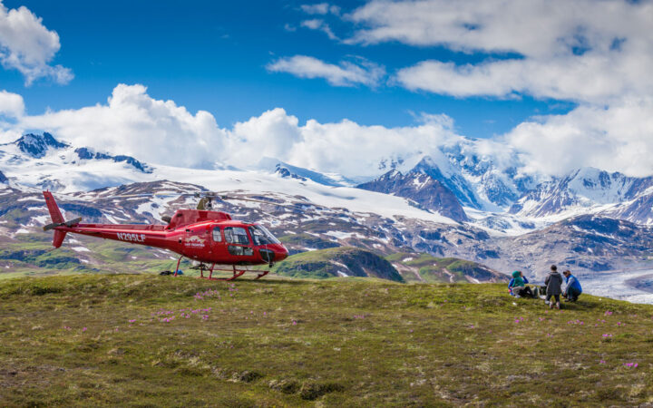 Red helicopter sits turned off atop a high meadow with snowy peaks beyond as Within the Wild Alaska guests have a picnic lunch.