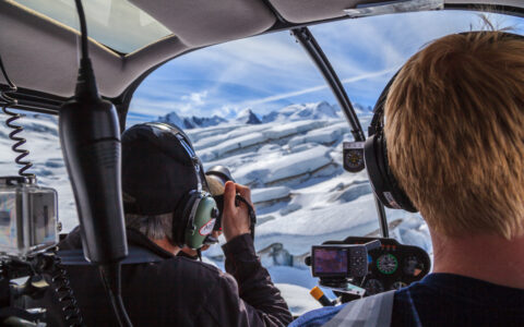 Man photographs glacier ice from the front of a helicopter while pilot flies on a sunny day during the Within the Wild Alaska tour.