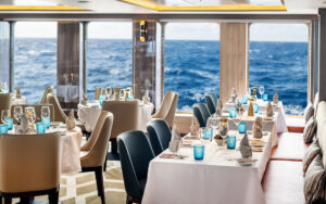 Dining room on World Voyager cruise ship with round & square 4-top tables set for dinner in white linens by large view windows.