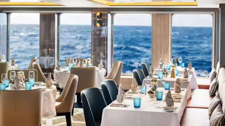 Dining room on World Voyager cruise ship with round & square 4-top tables set for dinner in white linens by large view windows.