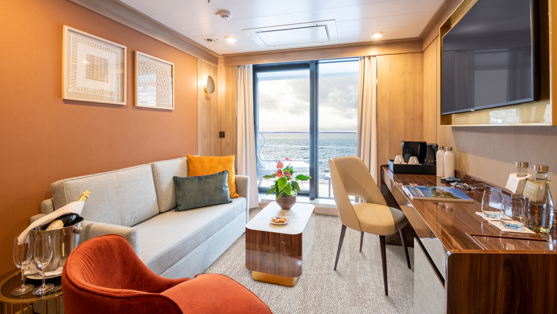 Second room of Deluxe Suite on World Voyager cruise ship with couch, coffee table, big desk, TV & sliding doors to balcony.