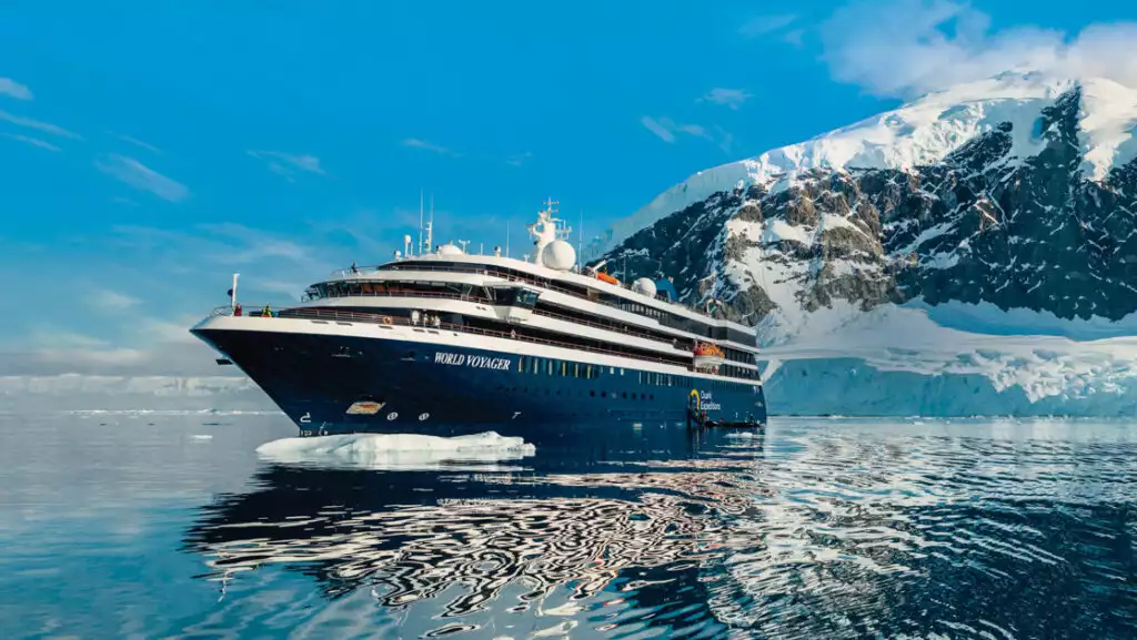 World Voyager cruise ship with blue hull & white upper decks sits in glassy Antarctic waters beside snowy peaks in the sun.