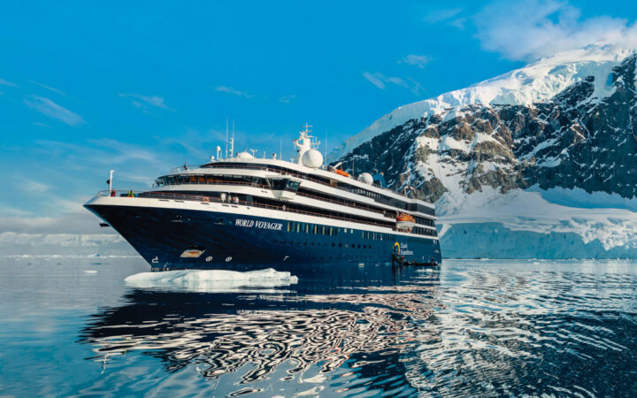 World Voyager cruise ship with blue hull & white upper decks sits in glassy Antarctic waters beside snowy peaks in the sun.