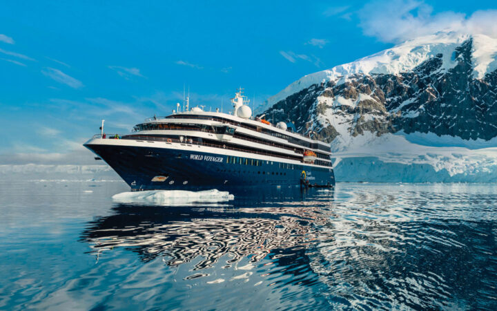 World Voyager cruise ship with blue hull & white upper decks sits in glassy Antarctic waters beside snowy peaks in the sun.