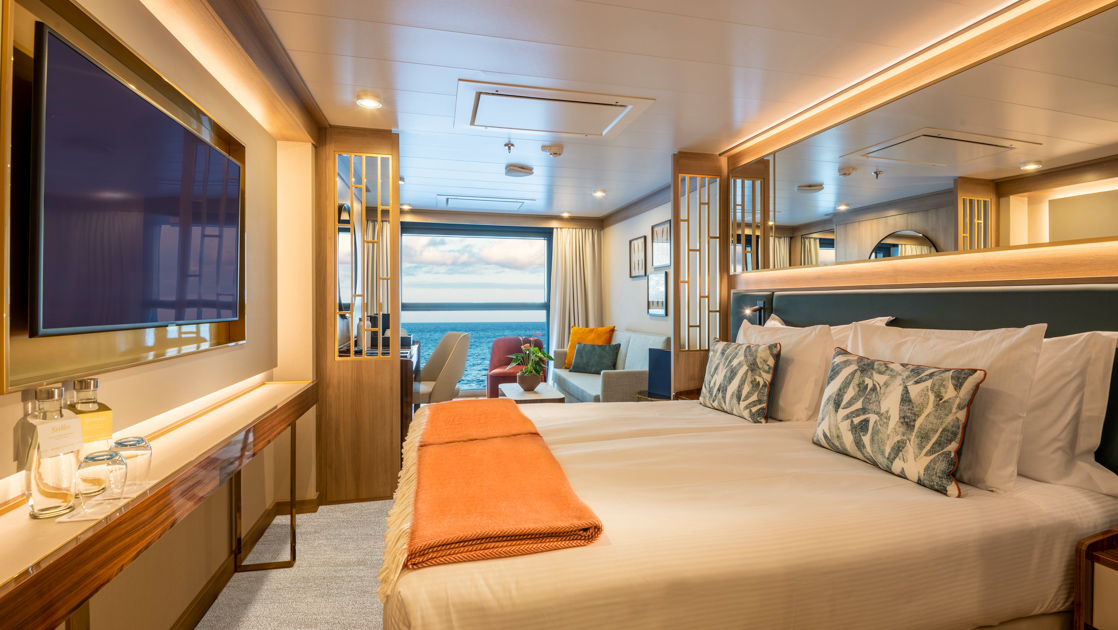 Infinity Suite on World Voyager cruise ship with double bed in white & peach linens, sitting area, desk & Juliette balcony.