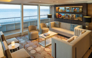 Observation Lounge on World Voyager cruise ship with modern beige couches & chairs, bookcase & floor-to-ceiling windows.