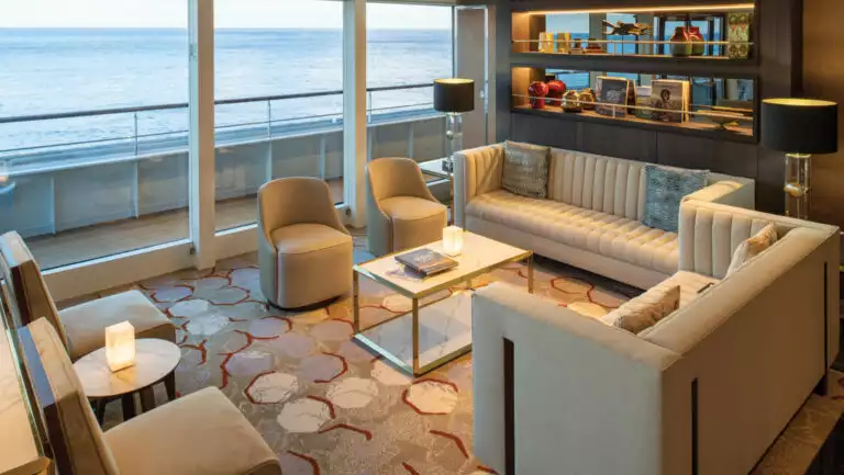 Observation Lounge on World Voyager cruise ship with modern beige couches & chairs, bookcase & floor-to-ceiling windows.