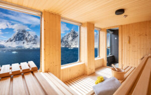 Sauna on World Voyager cruise ship with 2 rows of wooden bench seating, towels, water bucket & a wall of view windows.