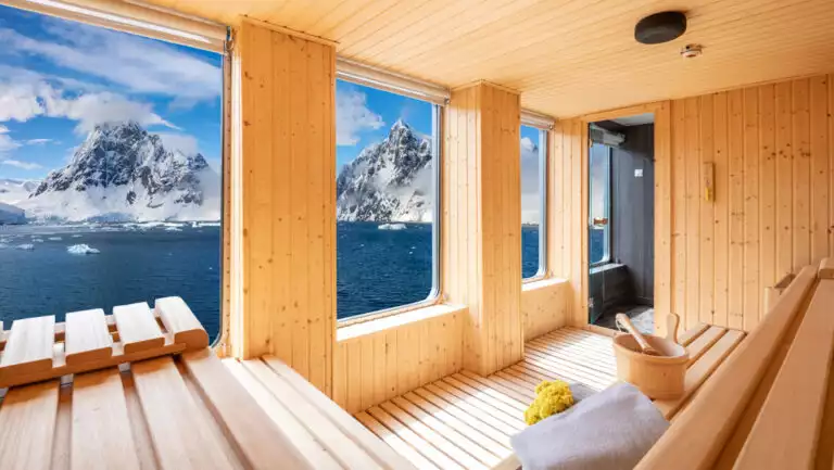 Sauna on World Voyager cruise ship with 2 rows of wooden bench seating, towels, water bucket & a wall of view windows.