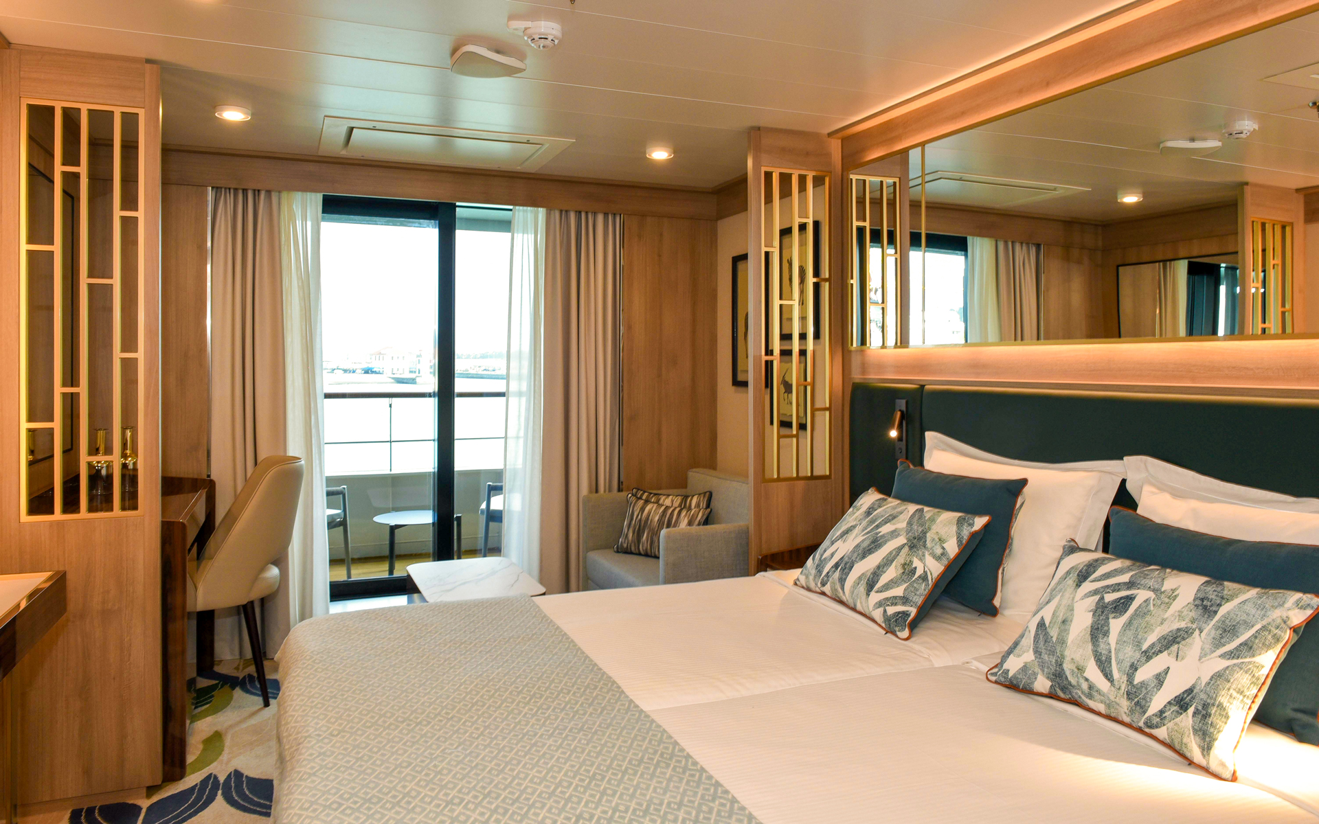 Veranda Suite on World Voyager cruise ship with double bed in white & green linens, sitting area, desk & step-out balcony.