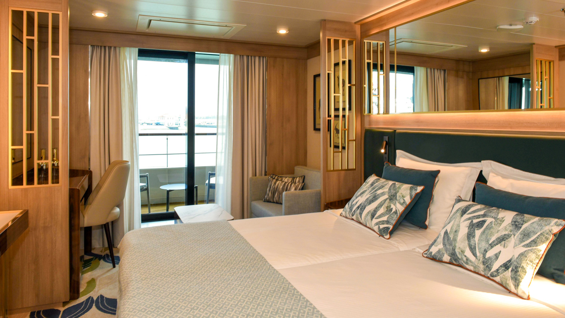 Veranda Suite on World Voyager cruise ship with double bed in white & green linens, sitting area, desk & step-out balcony.