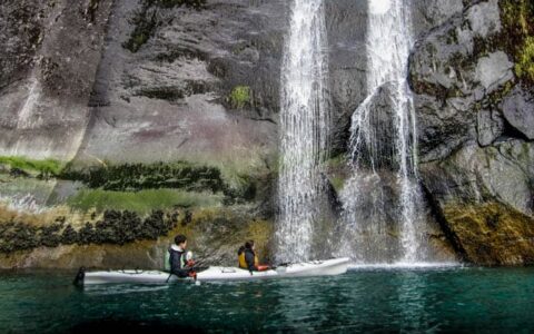 alaska traveler in a kayak on still water sits in front of a sheer rocky cliff with a waterfall coming down it