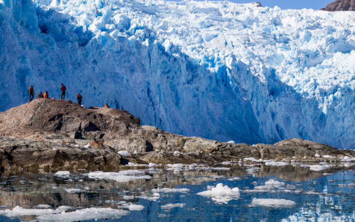 Several people standing on rocks in front of a massive blue and white glacier in Patagonia with ice floating all around them